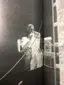 Black and white photograph of performer on stage captured through backstage rigging and equipment, demonstrating theater and live performance production technical setup